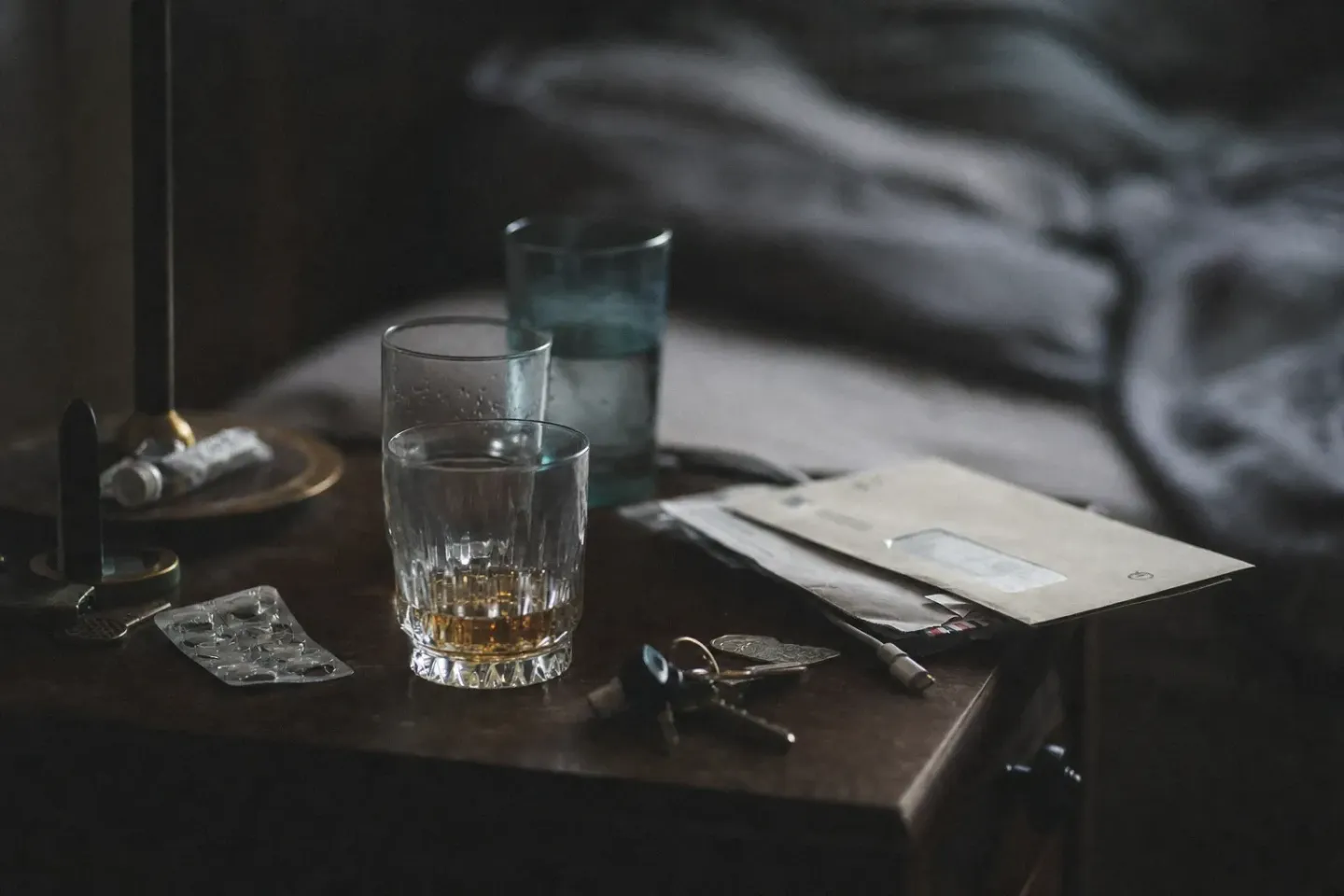 A cluttered bedside table in the morning: three half-drunk glasses, unopened post, an unplugged charger, a blister pack of medication and a set of keys, nobody in frame