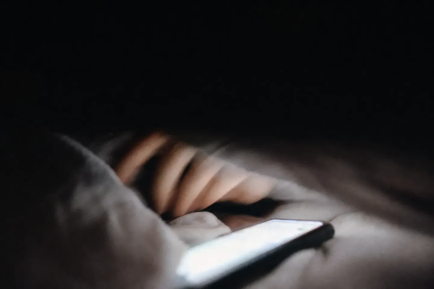 베개 높이에서 잡은 접사. 흔들린 손이 휴대폰에서 물러나 이불 속으로 되돌아가고, 화면은 읽을 수 없는 흰 얼룩으로 번져 있으며 나머지는 전부 어둠이다