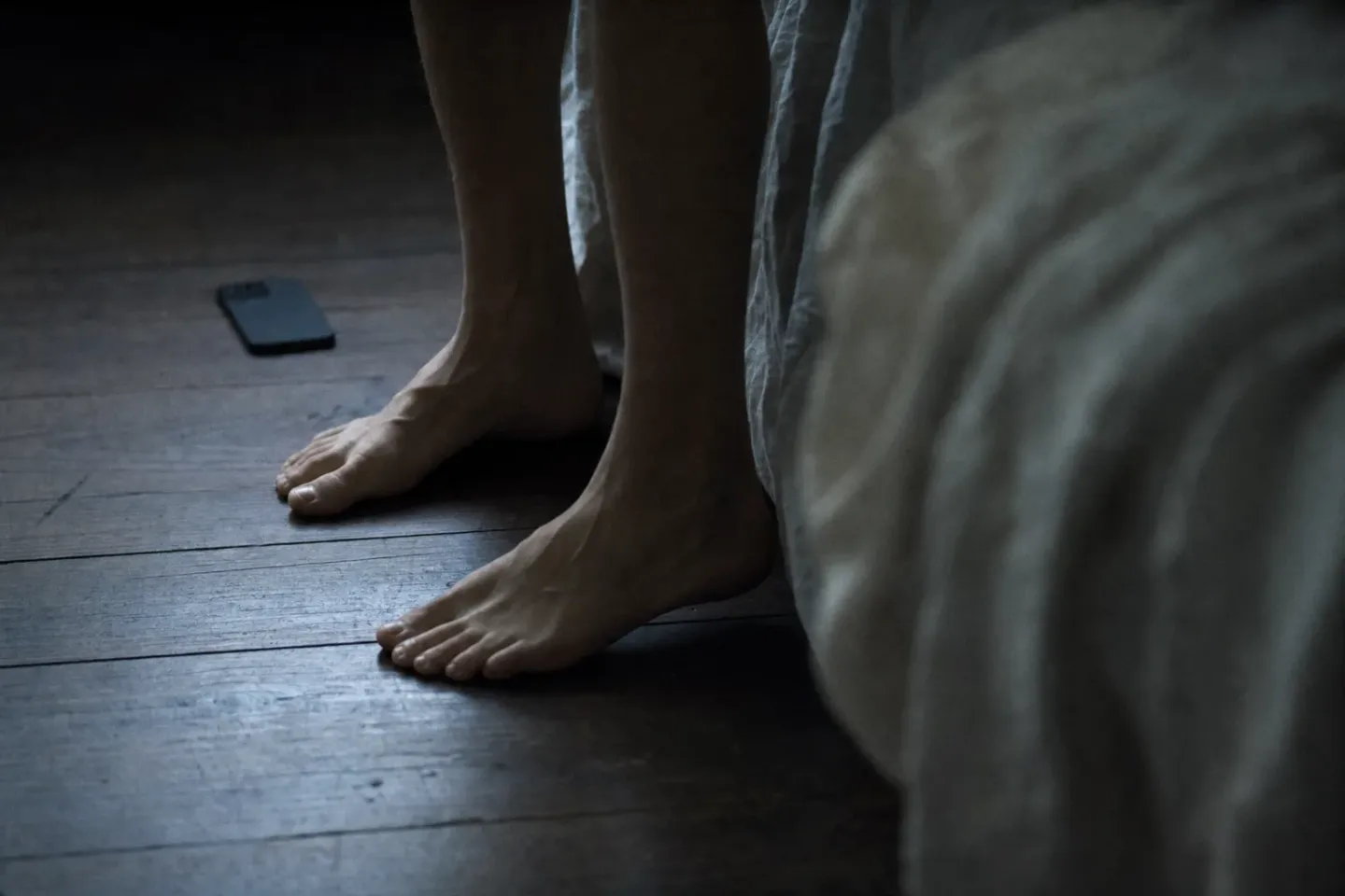 Bare feet planted on cold floorboards beside an unmade bed at dawn, a phone left lying face-down behind them