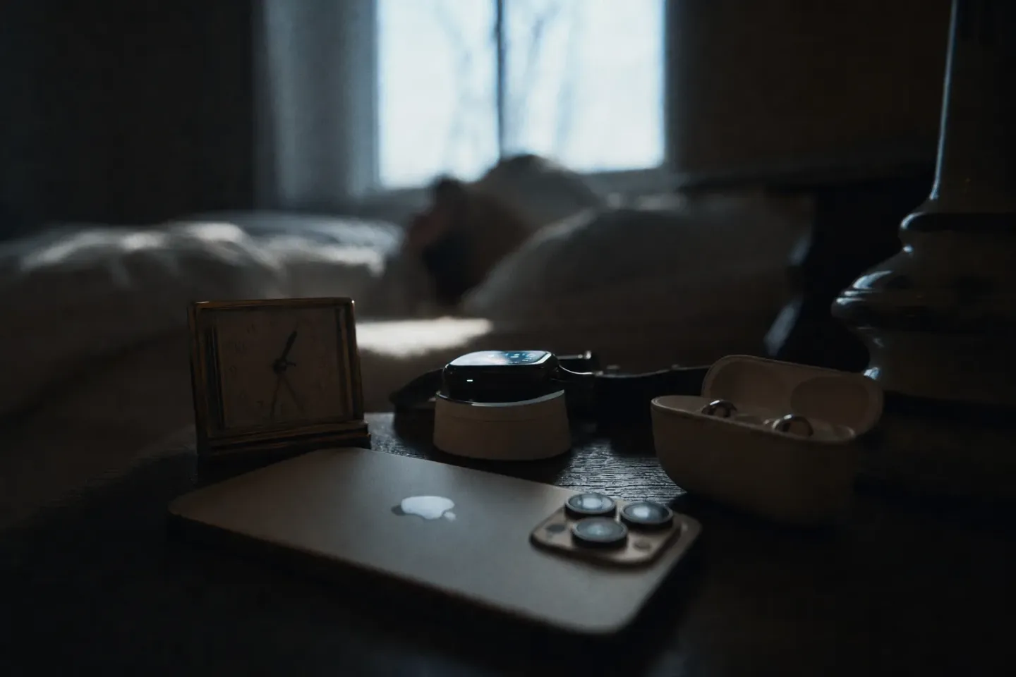 A cluttered bedside table at dawn holding several alarms at once: a face-down phone, a small travel clock and a smartwatch on its charger