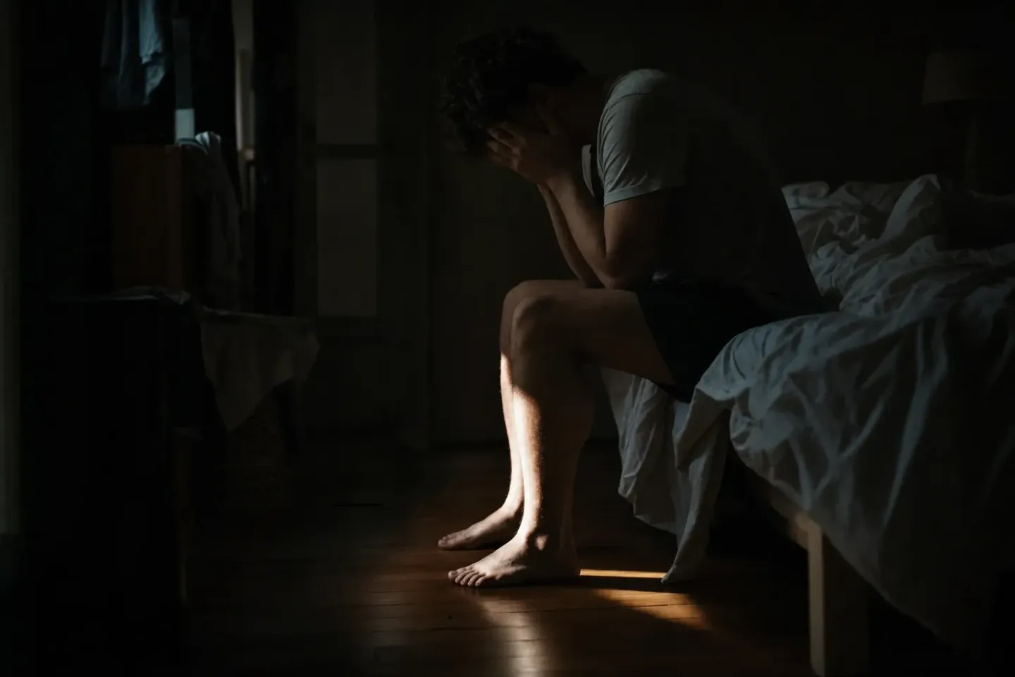 Une personne assise au bord d’un lit défait, tête entre les mains, une lame de lumière matinale coupant la pénombre de la chambre