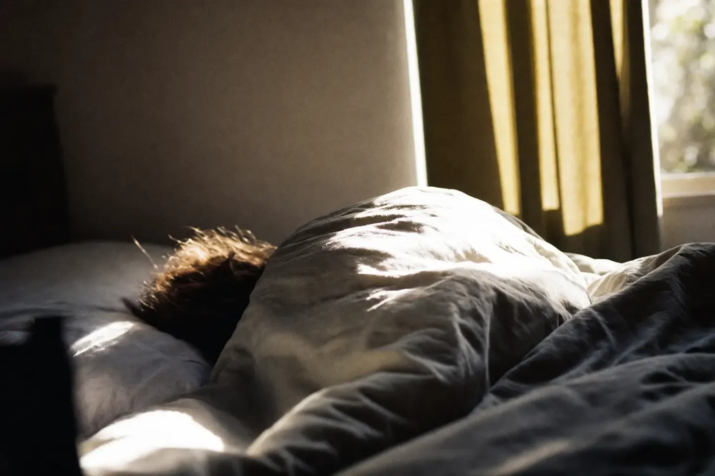 Soleil dur de fin de matinée tombant sur un lit encore occupé, draps froissés en pleine lumière