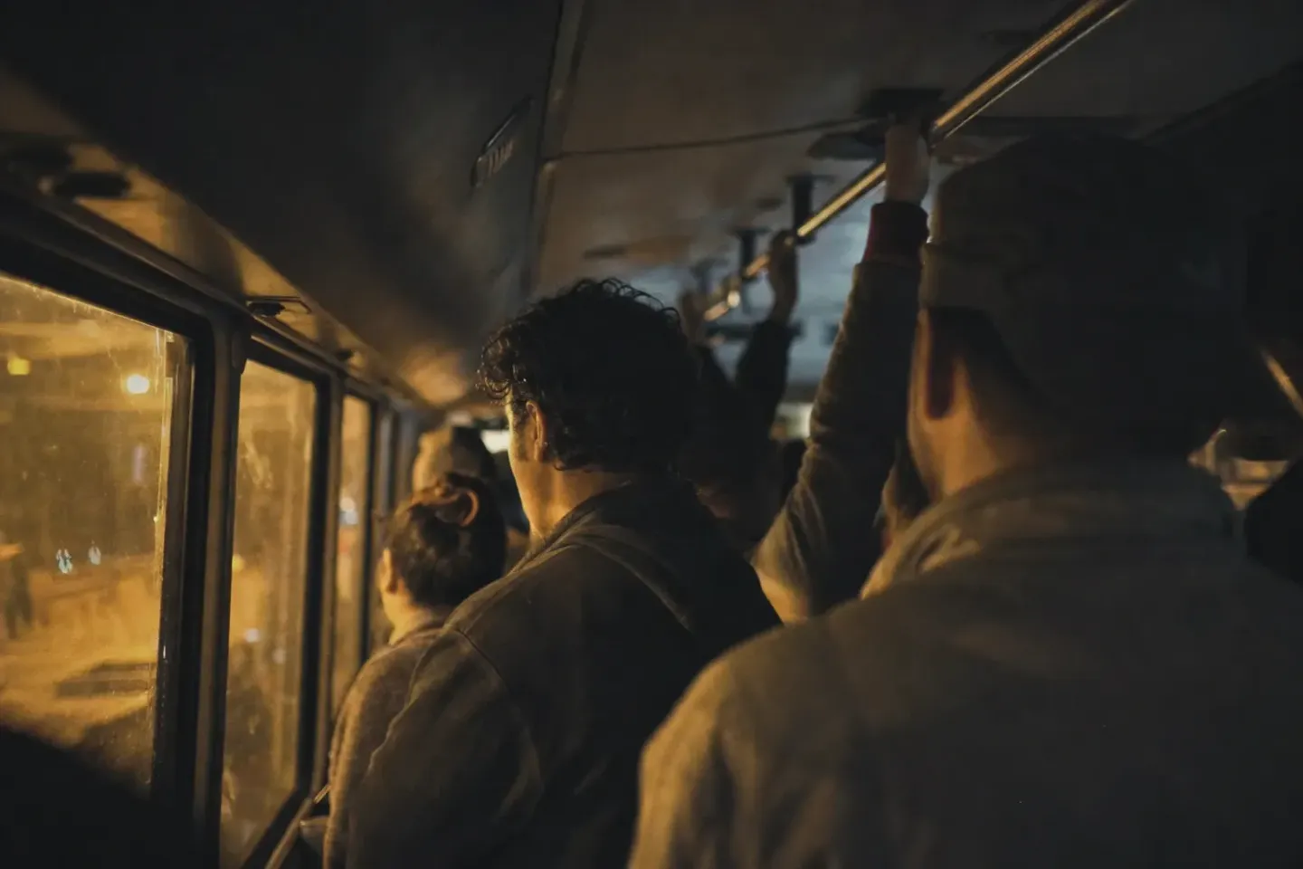 Interior de un microbús lleno antes del amanecer, con pasajeros de pie agarrados al tubo y la cara vuelta hacia las ventanas mientras pasa la luz de sodio de la avenida