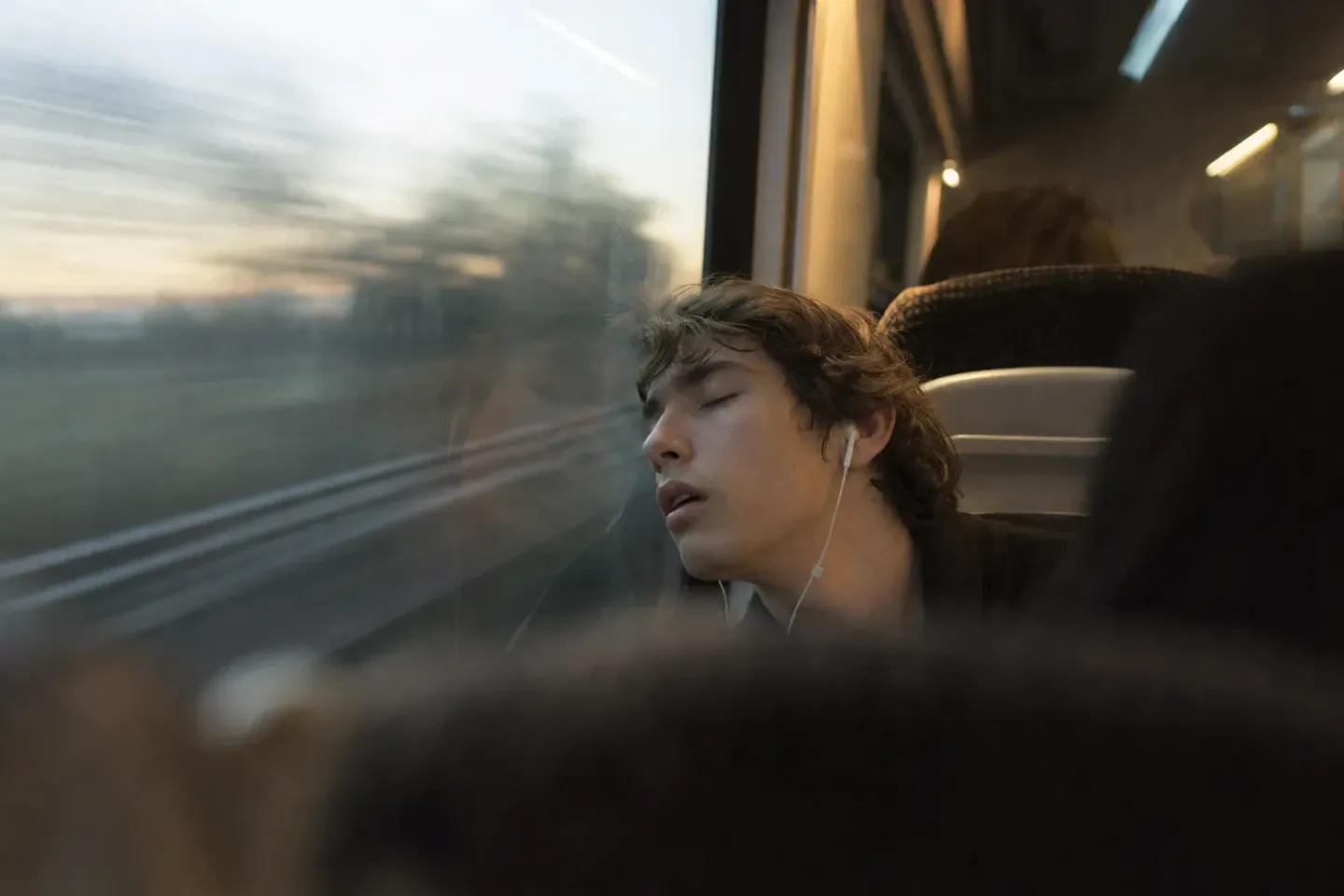 Un giovane passeggero addormentato accanto al finestrino di un regionale, la testa reclinata all'indietro e la bocca socchiusa, un auricolare a filo bianco ancora nell'orecchio, il paesaggio fuori ridotto a una scia orizzontale