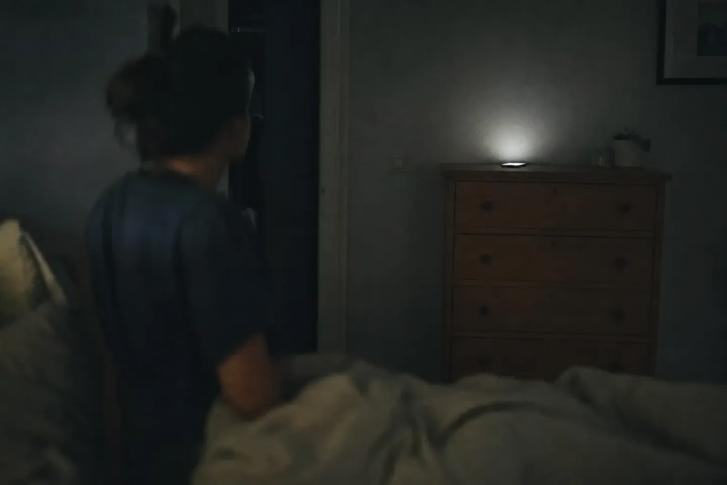 Alguien sentado en la cama, de espaldas y desenfocado en primer plano, mira hacia un teléfono encendido sobre una cómoda al otro extremo del dormitorio