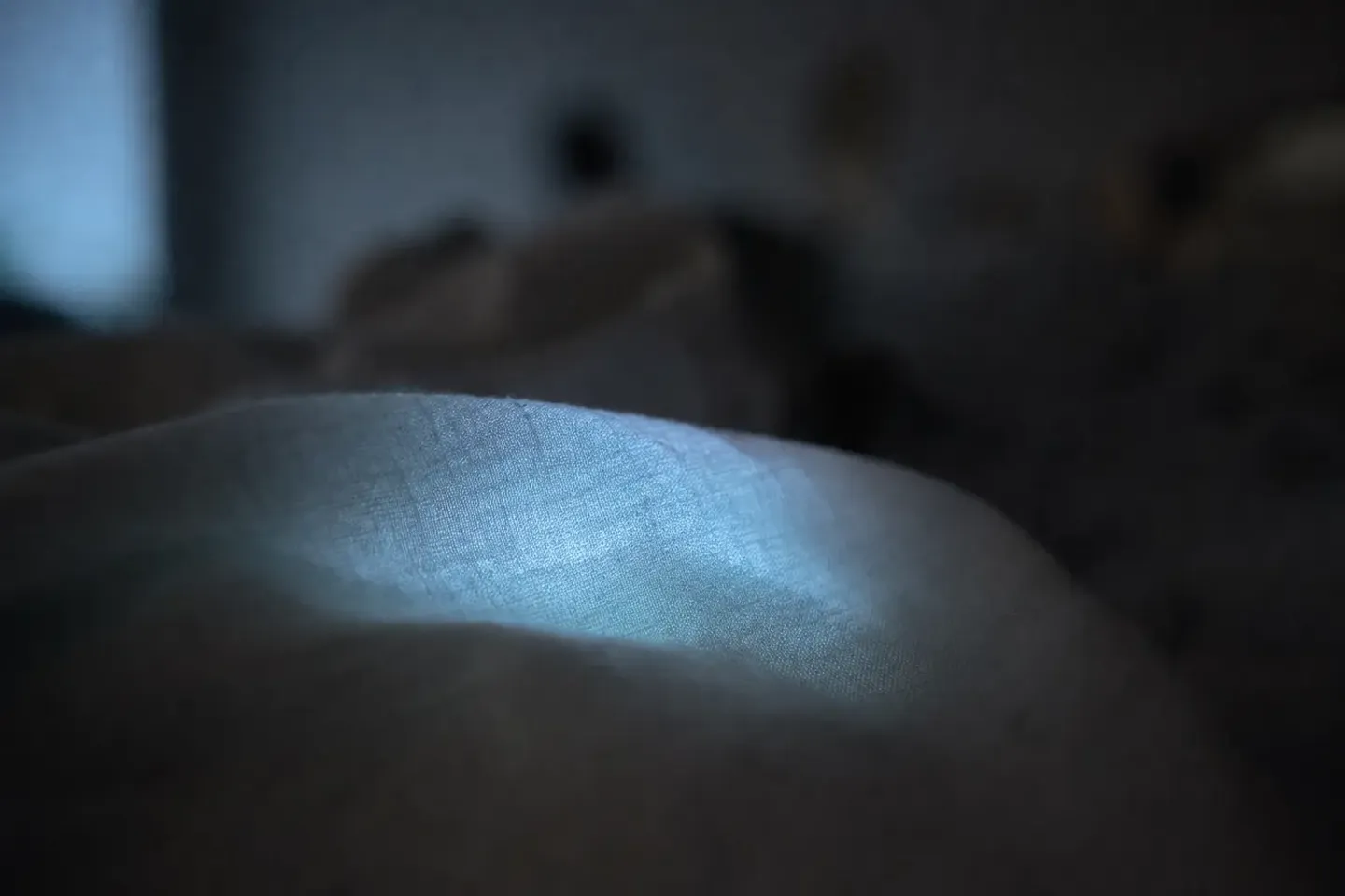 Koud telefoonlicht schijnt gedempt door de plooien van een dekbed; het toestel zelf is niet te zien