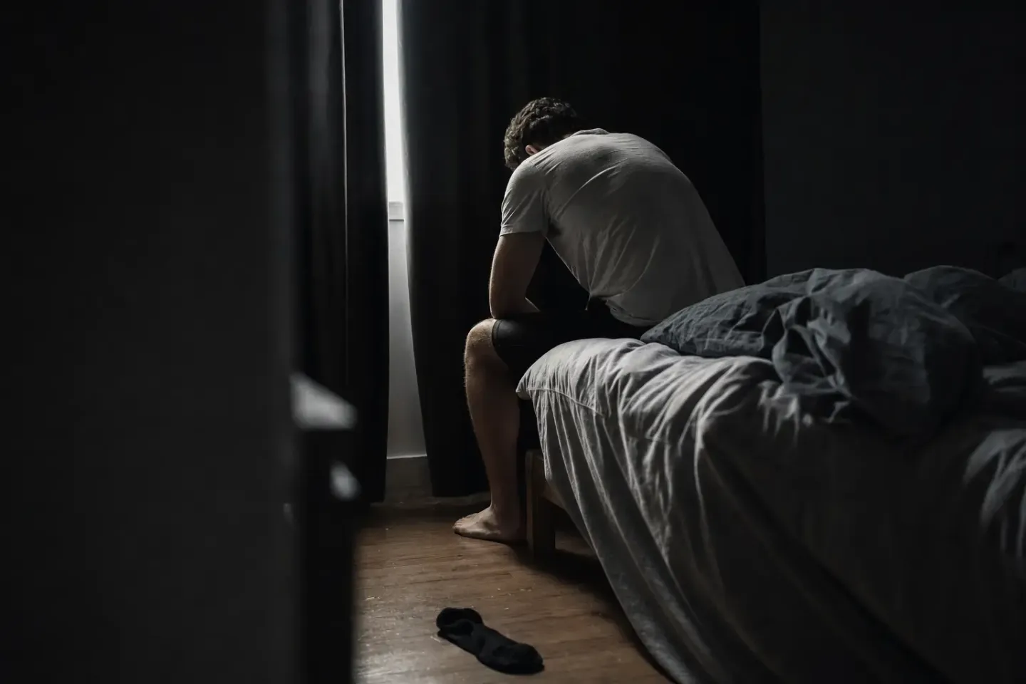 Iemand zit bij het eerste ochtendlicht roerloos op de rand van een onopgemaakt bed, voeten op een koude vloer