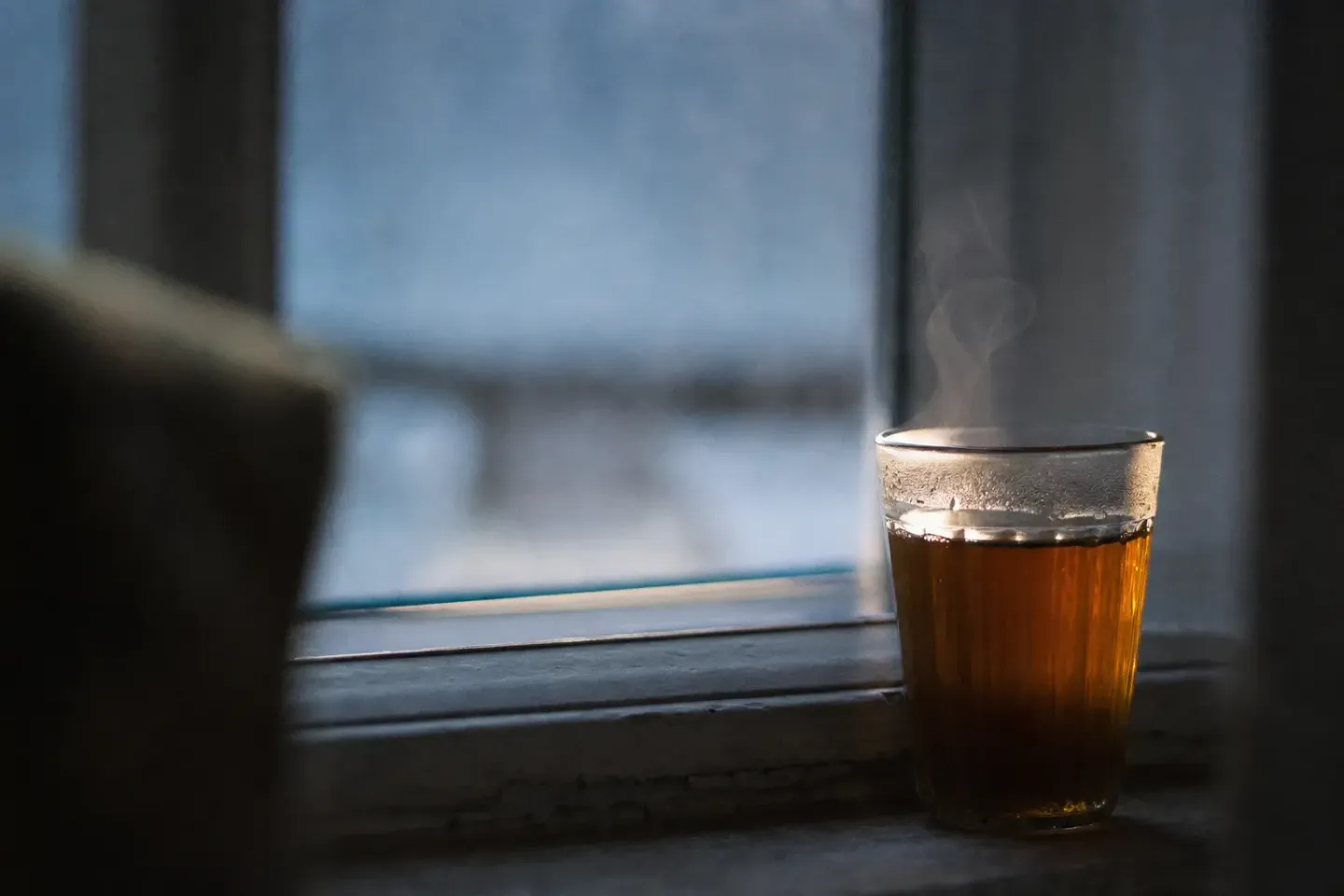 Un verre de thé posé sur un rebord de fenêtre avant l’aube, la vapeur captant la seule lumière de la pièce