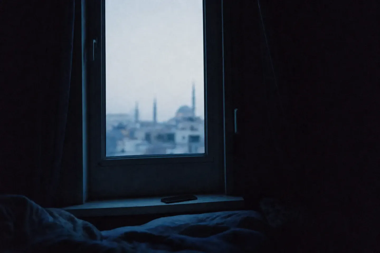 Vue depuis un lit dans une chambre encore sombre : la fenêtre forme un rectangle pâle où l’on devine à peine des toits, un téléphone posé face contre l’appui
