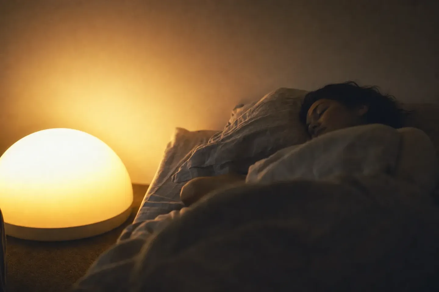 Le même simulateur d’aube à pleine intensité, tout près du lit ; à côté, une dormeuse immobile sur l’oreiller, les yeux fermés et le visage détendu, le reste de la chambre encore dans le noir