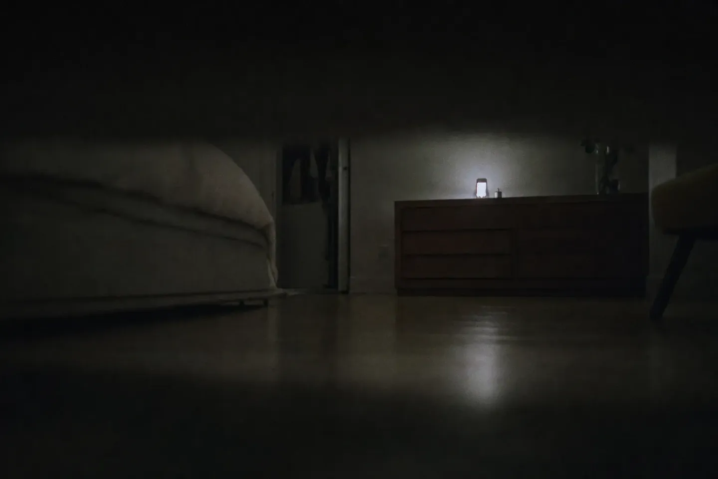 A bedroom seen from floor level under a duvet, with a lit phone on a chest of drawers at the far end of the room and the whole width of the floorboards in between
