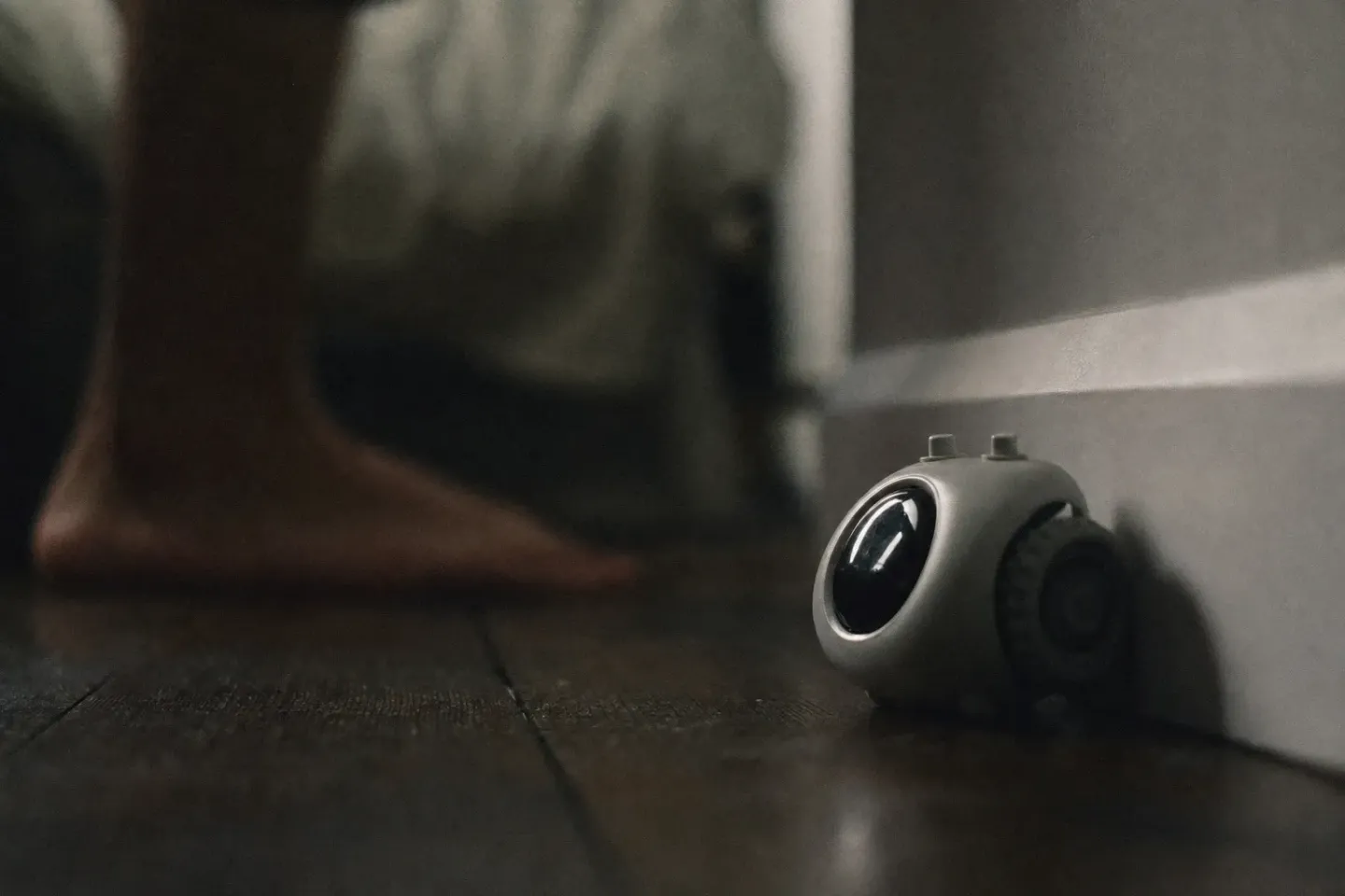 A small wheeled alarm clock jammed against a skirting board in a dark bedroom, seen from floor level, with a blurred bare foot behind it