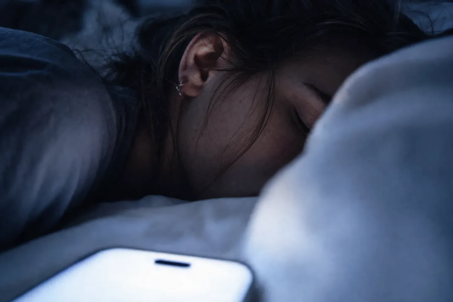 Gros plan sur l’oreille d’une jeune femme endormie, le visage dans l’oreiller, tandis que l’écran allumé de son téléphone brille à vingt centimètres dans le flou du premier plan
