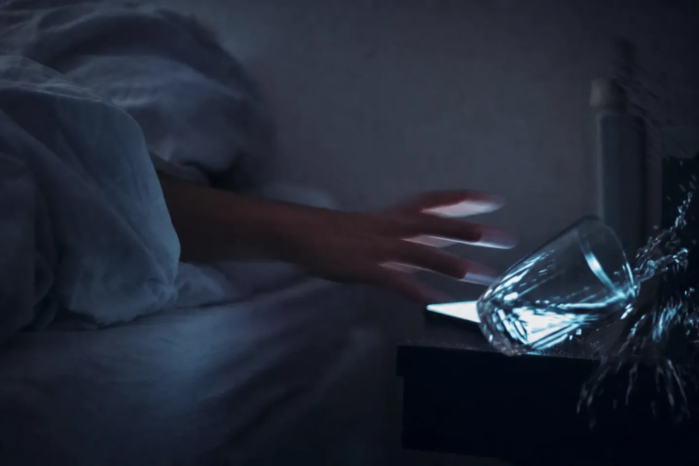 Une main sortie de sous la couette cherche à tâtons dans le noir, en flou de bougé, et renverse un verre d’eau à côté d’un téléphone allumé