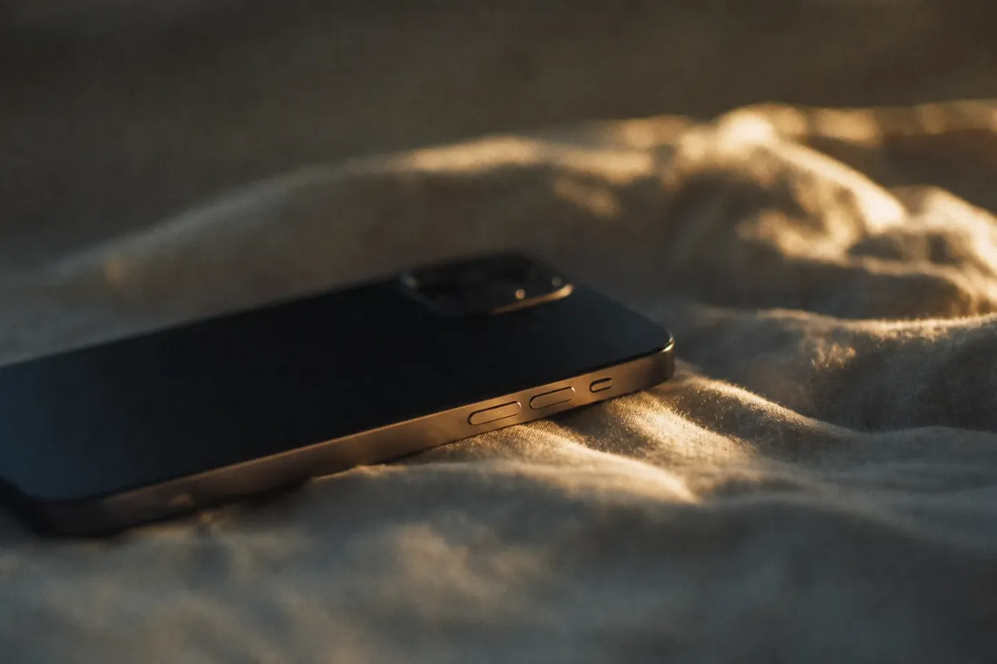 Altın rengi sabah ışığında, çarşafın üzerinde yüzüstü duran bir iPhone’un yan ses tuşları yakın planda