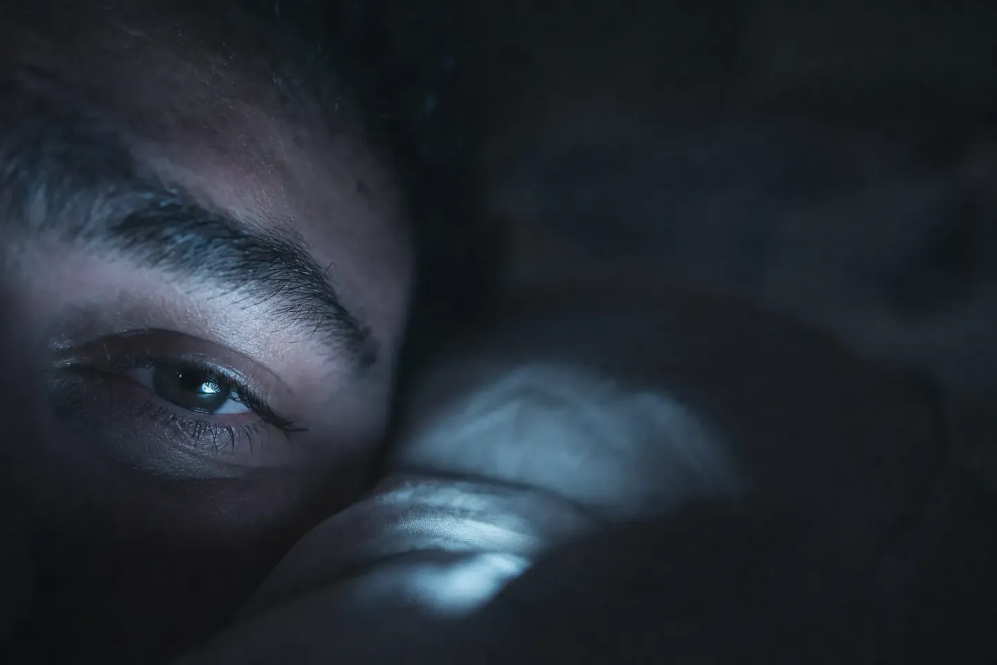 Extreme close-up of a half-open eye pressed into a pillow in the dark, lit by the cold glow of a phone out of frame