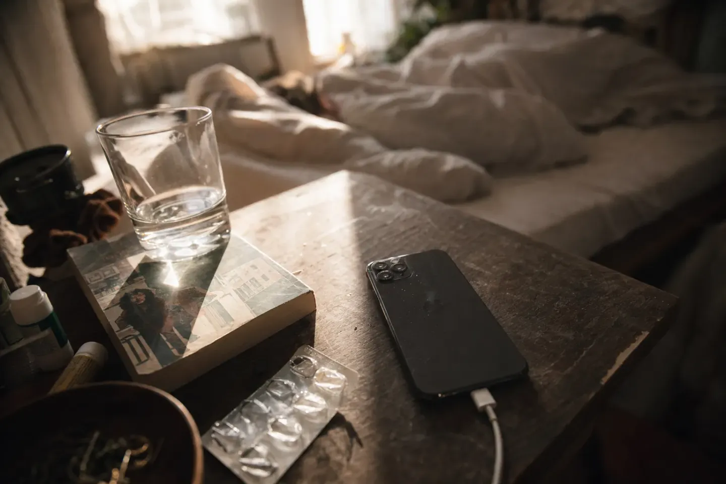 A phone lying face-down on a cluttered nightstand in hard late-morning daylight, a bed still occupied behind it