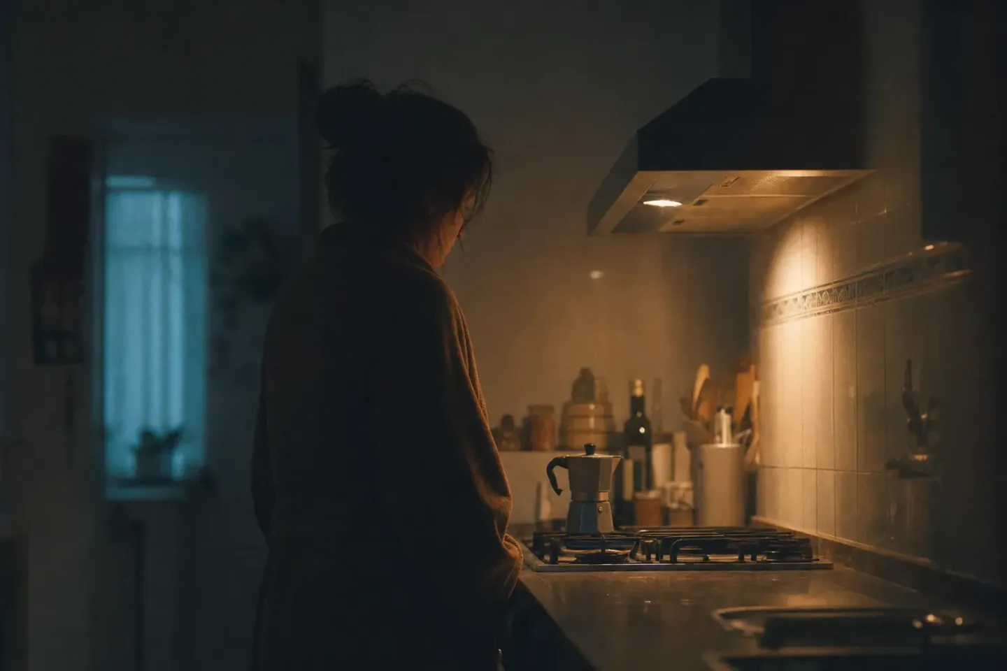 Una persona en bata, quieta delante de una cafetera italiana en una cocina española antes de que salga el sol, iluminada solo por la llama del gas