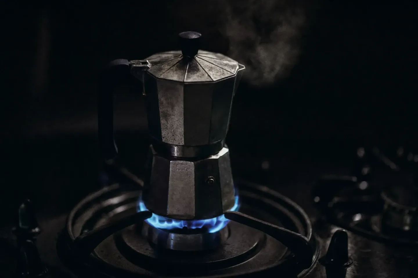 Primer plano de una cafetera italiana sobre el fuego en una cocina a oscuras, con la llama azul del gas como única fuente de luz