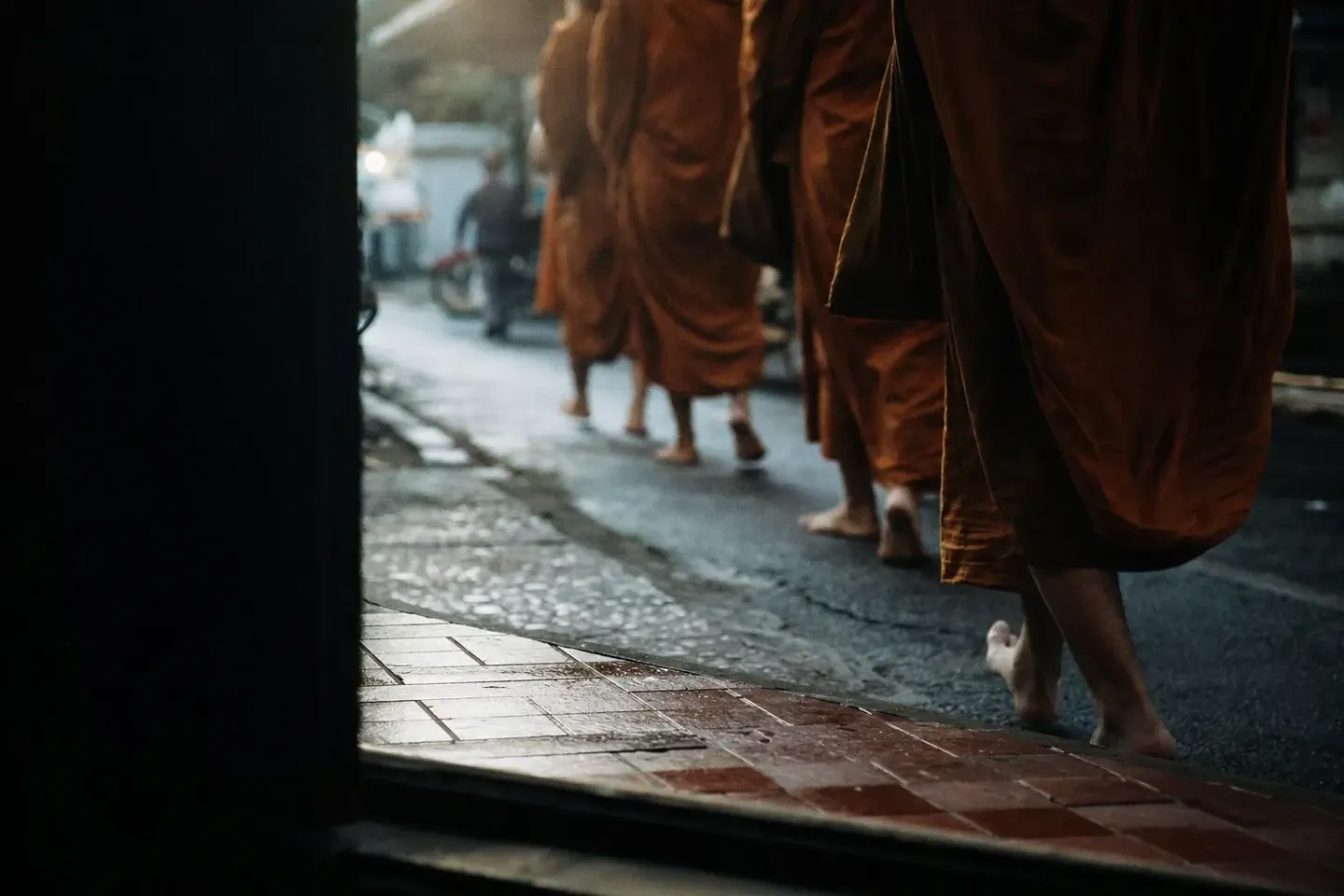 ถนนในซอยของไทยตอนฟ้าเพิ่งสาง มองจากธรณีประตูเตี้ย ๆ มีแถวผ้าสีเหลืองส้มเดินผ่านไป พื้นถนนยังเปียก และไม่เห็นใบหน้าใครในภาพ