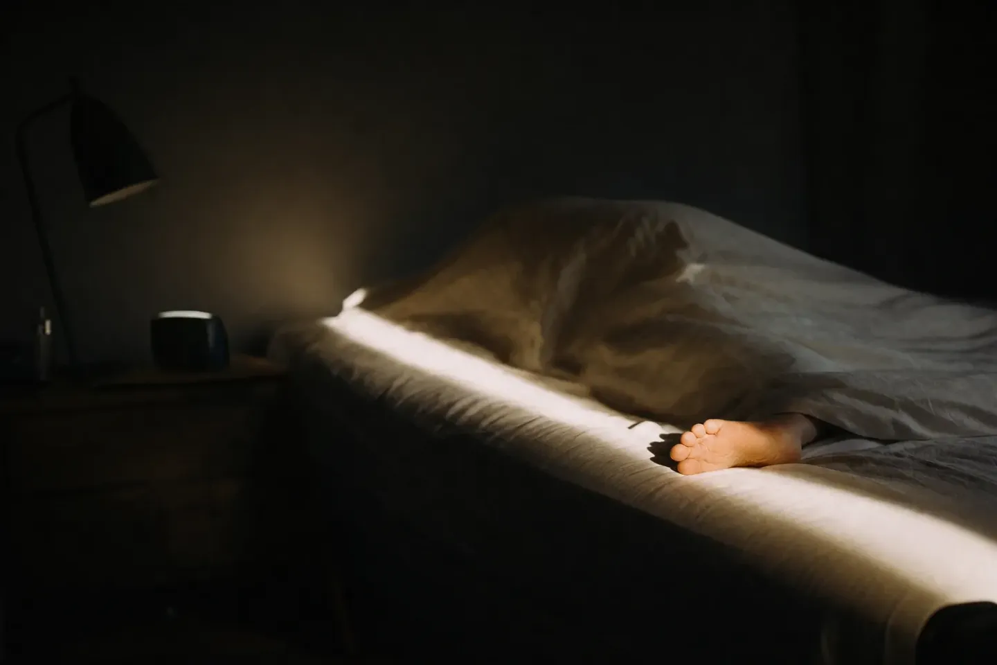 Dunkles Schlafzimmer: ein greller Lichtstreifen fällt quer über das Bett, unter der Decke liegt formlos eine Person, nur ein nackter Fuß schaut heraus; auf dem Nachttisch stehen eine Leselampe und ein kleines dunkles Gerät, dessen Anzeige nicht zu sehen ist, alles außerhalb des Streifens liegt in tiefem Schatten
