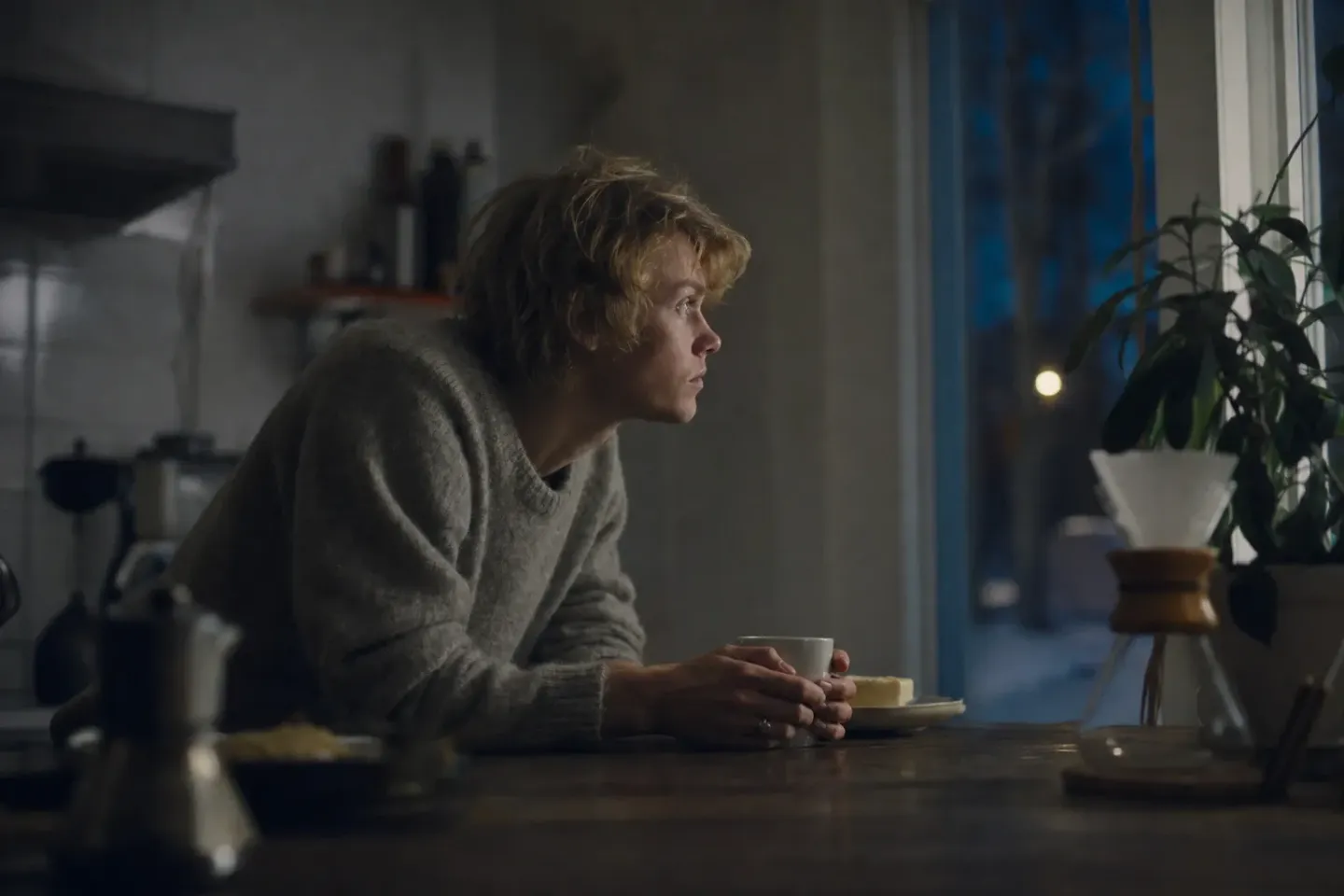 Ett svenskt kök klockan sju en vintermorgon där någon lutar sig med båda underarmarna mot bänken över en mugg och stirrar rakt fram medan det fortfarande är mörkt ute