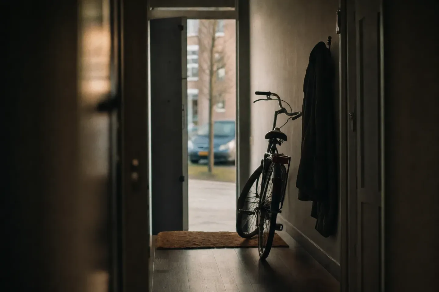 Een Nederlandse gang om acht uur 's ochtends: een fiets tegen de muur, een jas half van de kapstok gegleden, de voordeur op een kier met grijs daglicht erachter, niemand in beeld