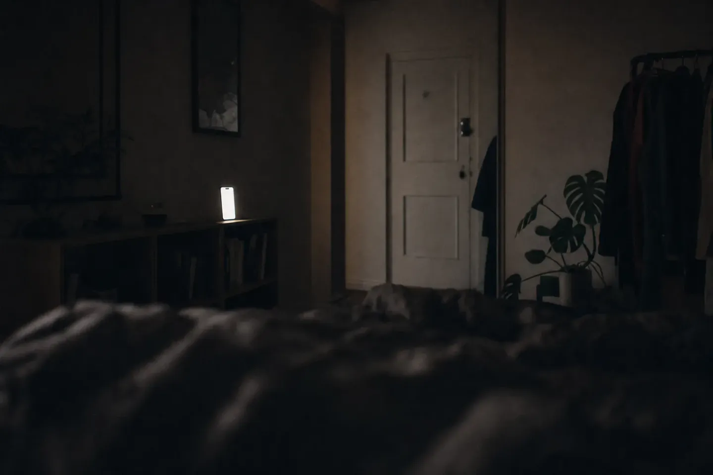 Blick aus dem Bett in ein dunkles Schlafzimmer, auf einem Regal an der gegenüberliegenden Wand leuchtet ein Telefon