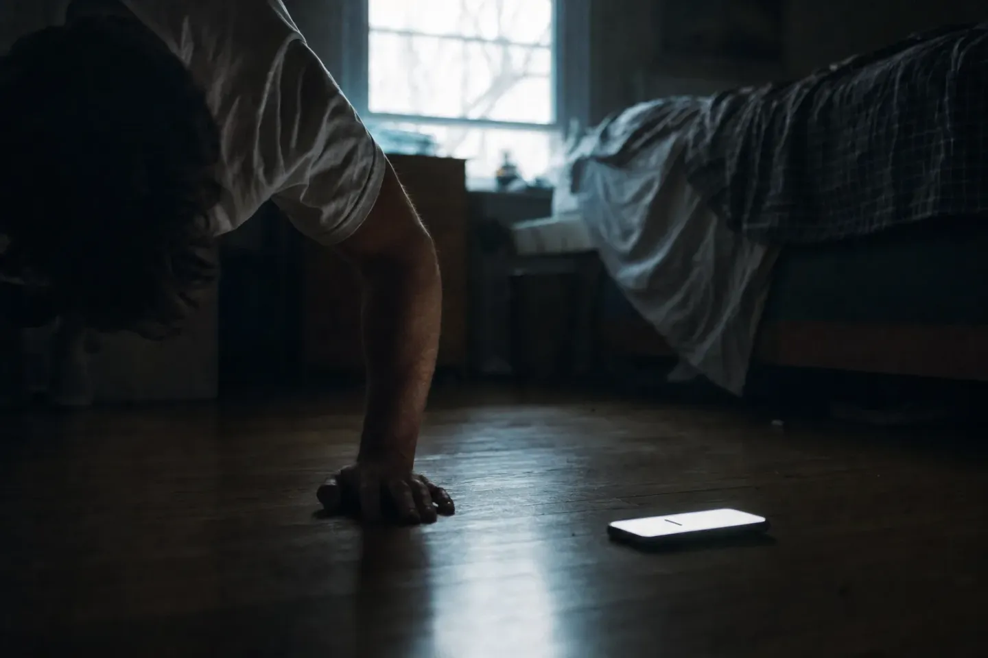 Jemand macht bei Morgendämmerung Liegestütze auf dem Fußboden neben einem zerwühlten Bett, das Telefon liegt vor den Händen