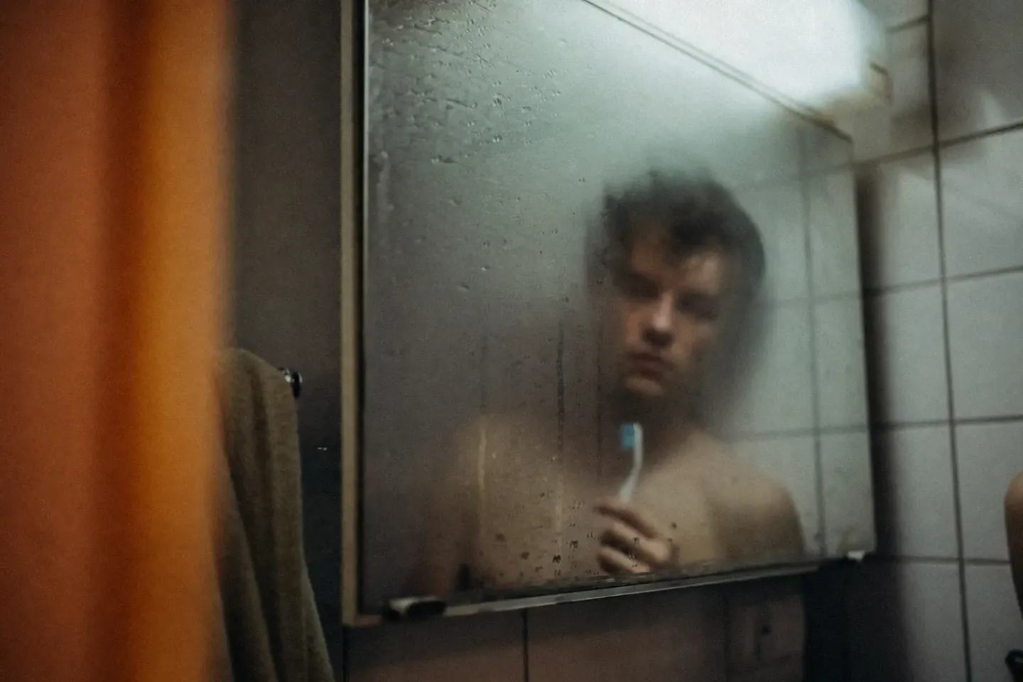 Een klein badkamertje: in een halfbeslagen spiegel kijkt een man met blote bovenlijf en warrig haar zijn eigen spiegelbeeld recht aan, uitdrukkingsloos, met een tandenborstel in zijn hand die hij niet gebruikt; links in beeld een oranje gordijn