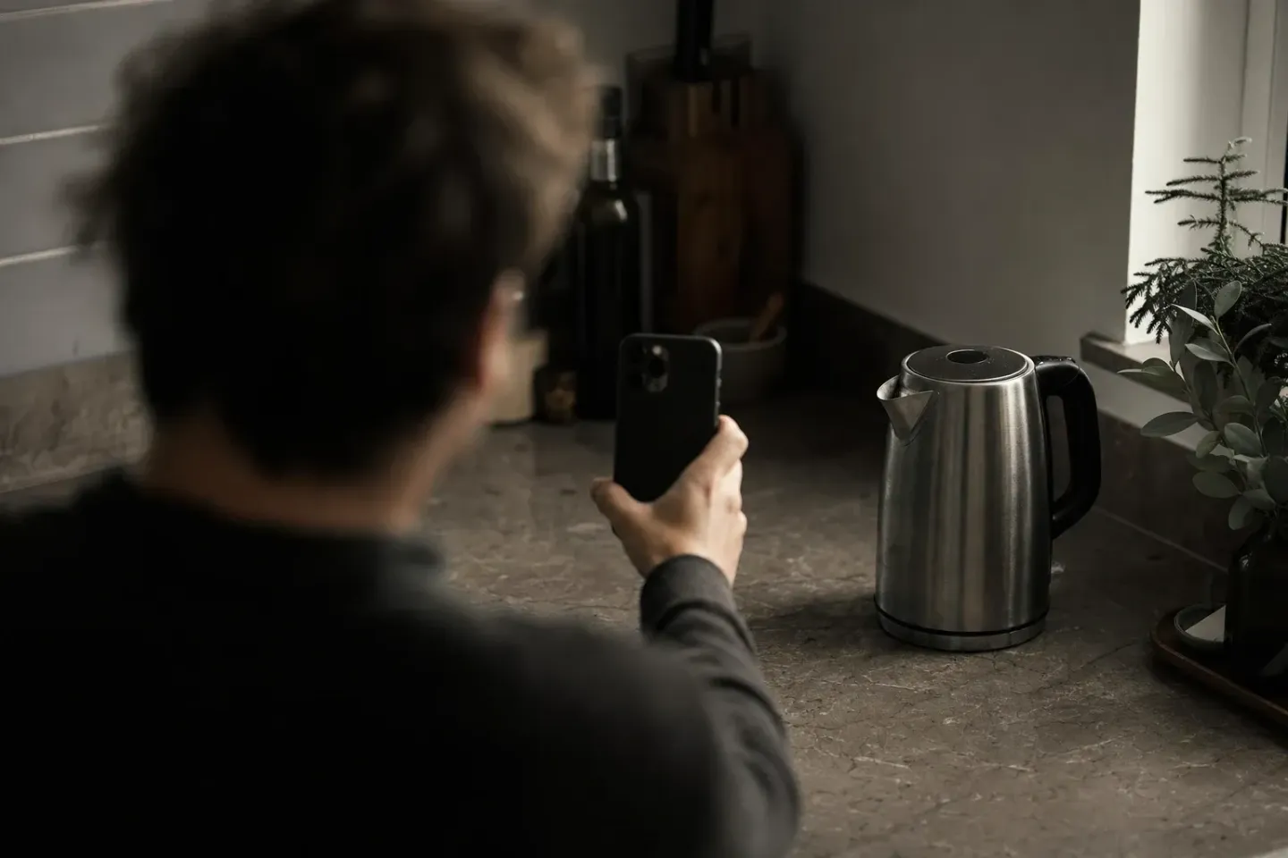 Seen over a blurred shoulder, someone points the back camera of a phone at a stainless-steel kettle standing in focus on a dim kitchen counter
