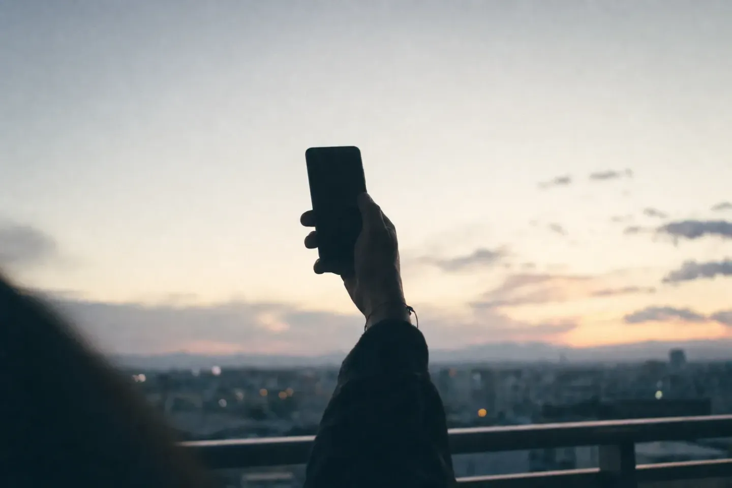 A silhouetted arm holding a phone up against a pale dawn sky, with a balcony rail and the lights of a waking city below, the screen turned away and unreadable