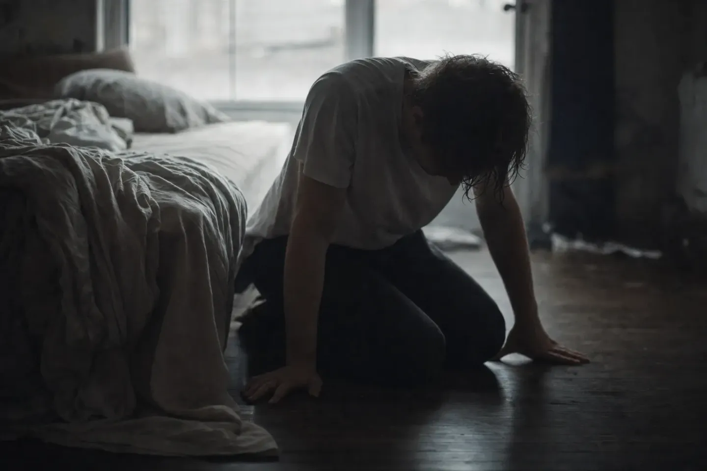 A man on his knees on the floorboards beside an unmade bed, head hung low, damp hair falling over his face, both palms flat on the boards, in flat grey backlight from the window