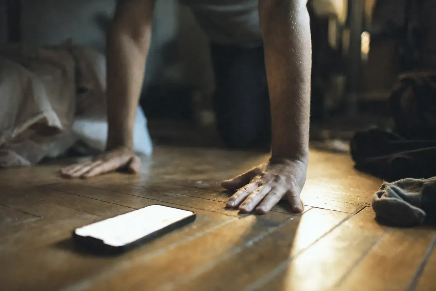 Ground-level macro of two hands flat on worn floorboards, forearms braced, the rest of the body out of focus above, a phone lying screen-up at the edge of frame in the raking light of dawn