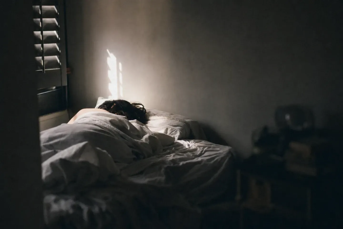 Luz cruda de lunes entrando por una persiana entreabierta sobre una cama todavía ocupada, con la persona dormida de espaldas a la luz