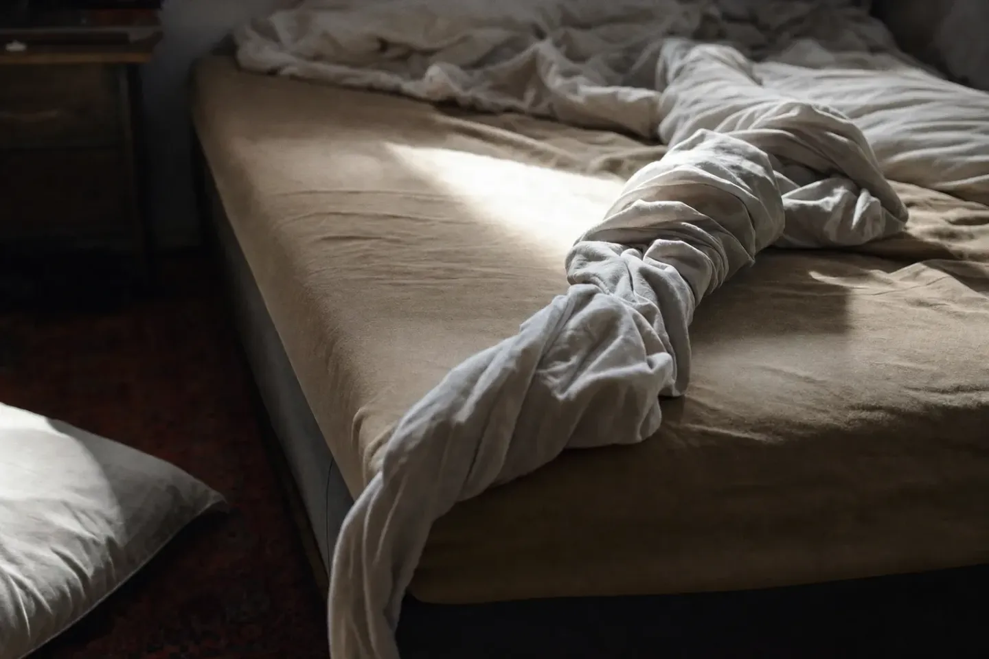 Detalle de una cama a media mañana con las sábanas retorcidas como una cuerda y una almohada caída en el suelo, con luz dura marcando los pliegues