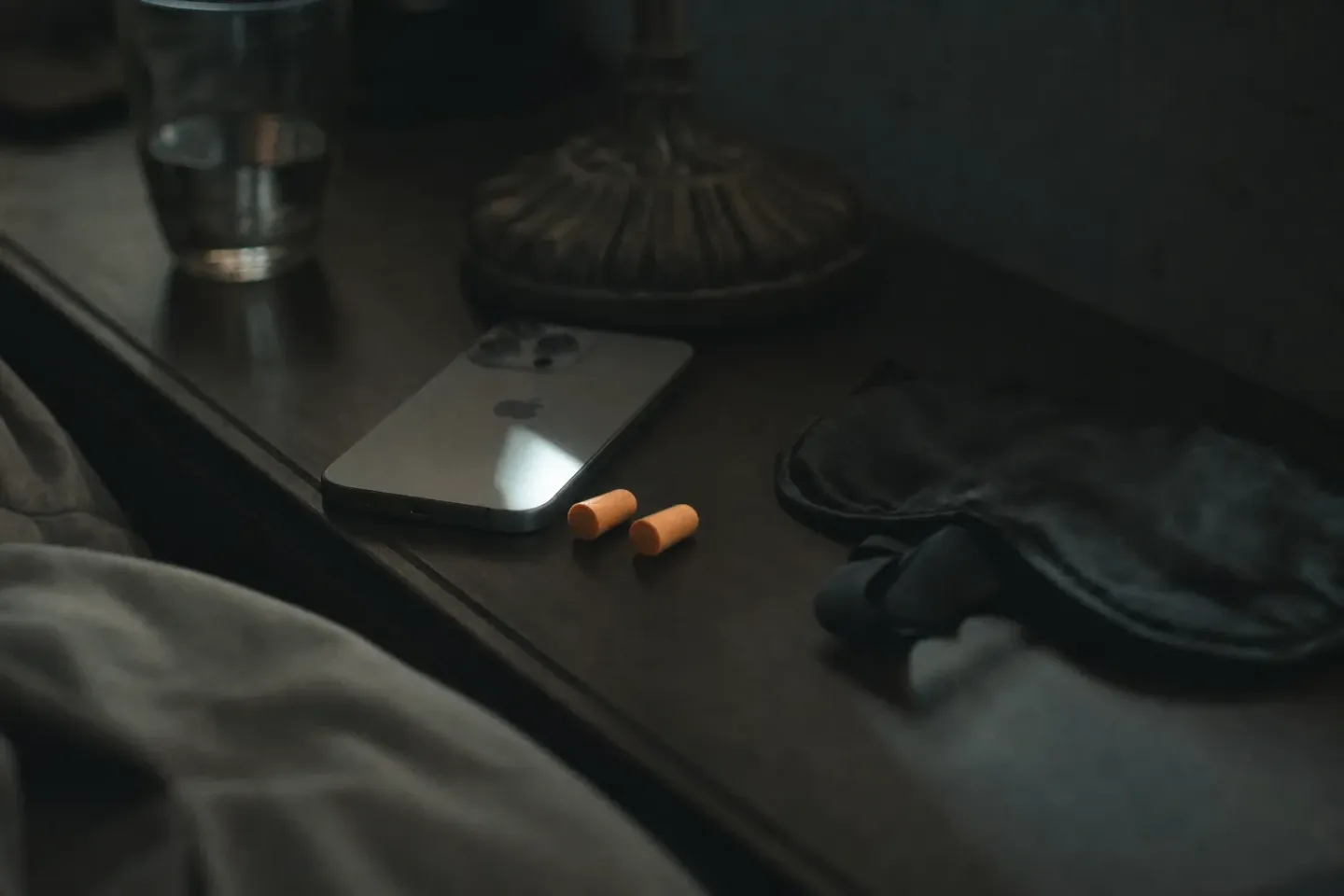 Een nachtkastje in het donker met een telefoon op zijn scherm, oordoppen van schuim en een slaapmasker ernaast