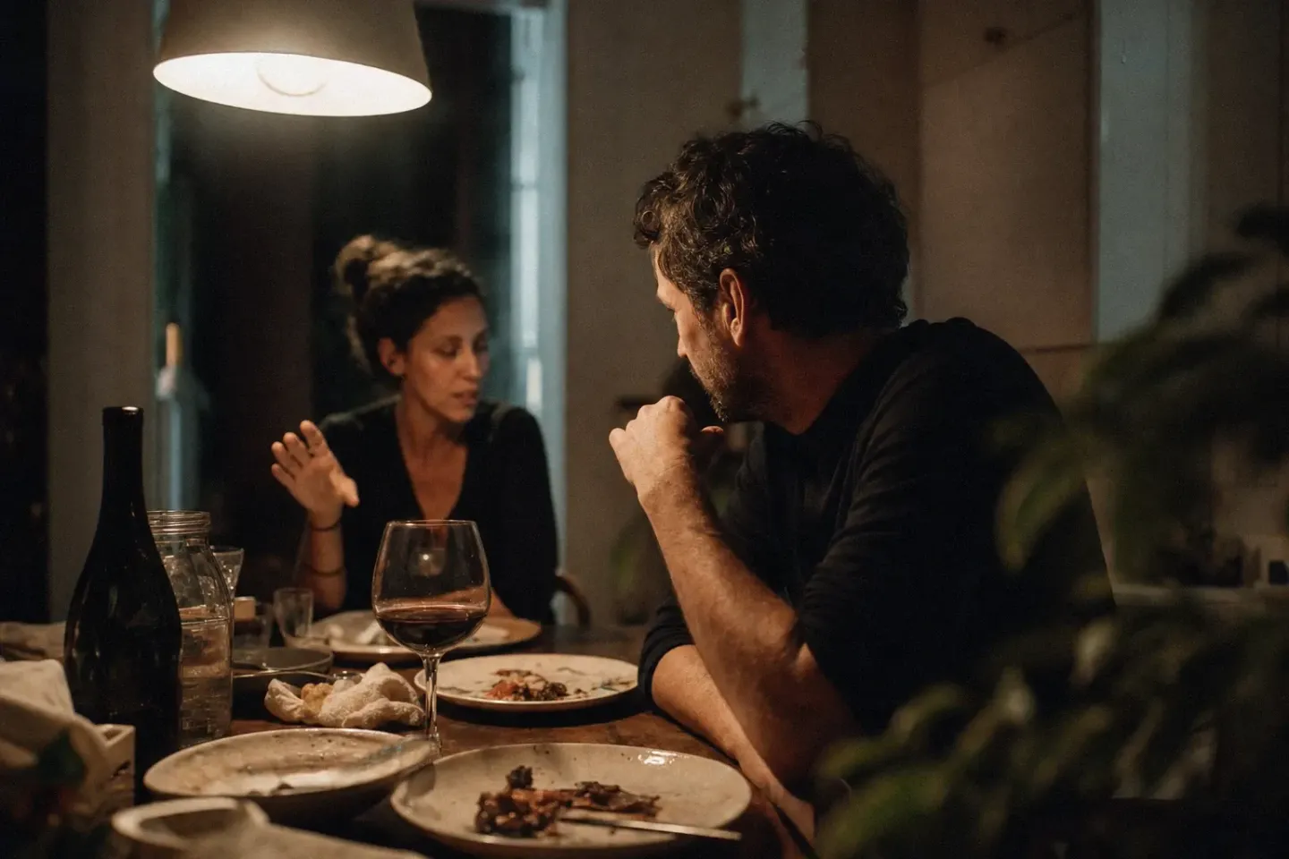 Sobremesa de noche bajo una lámpara de techo: platos a medio recoger, una botella vacía y una copa de vino tinto, mientras dos personas siguen hablando en la mesa