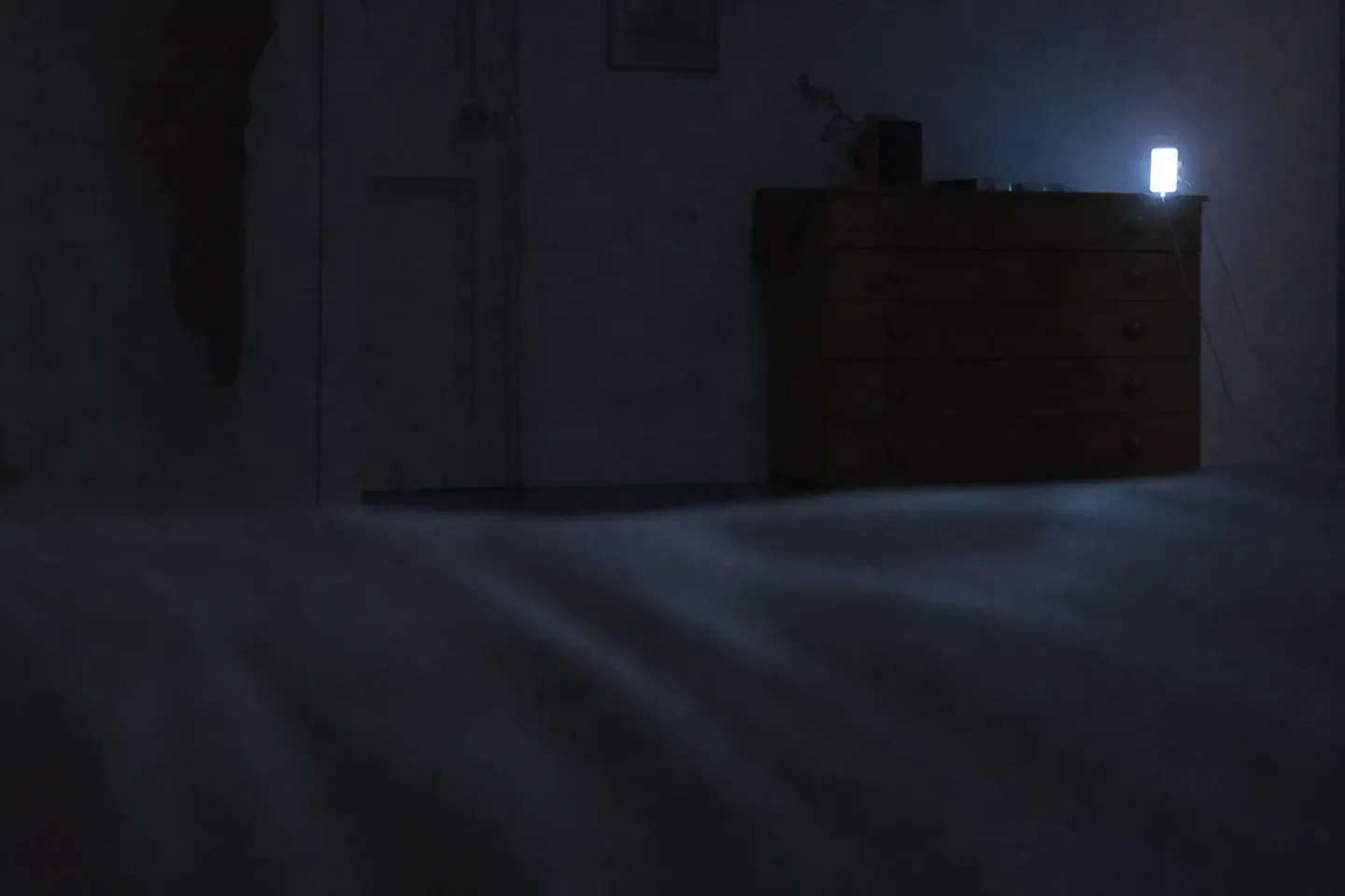 Seen from the pillow across a dark bedroom, a phone glowing on a chest of drawers against the far wall