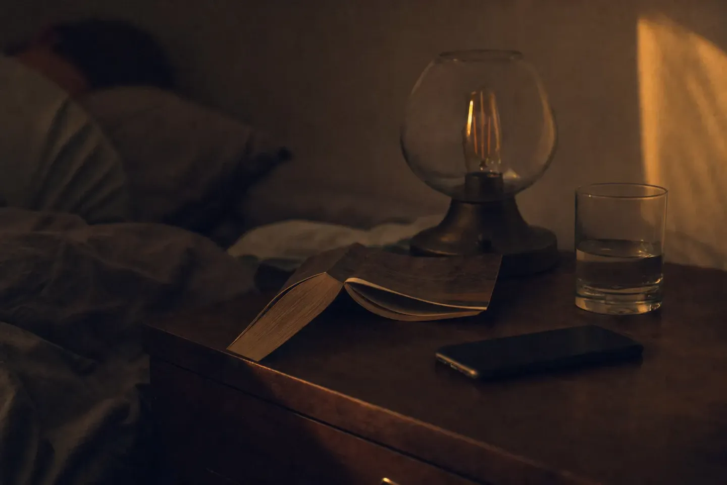 A bedside table at night: a lamp still lit, a paperback face-down, a glass of water, the phone pushed away face-down, someone already asleep behind