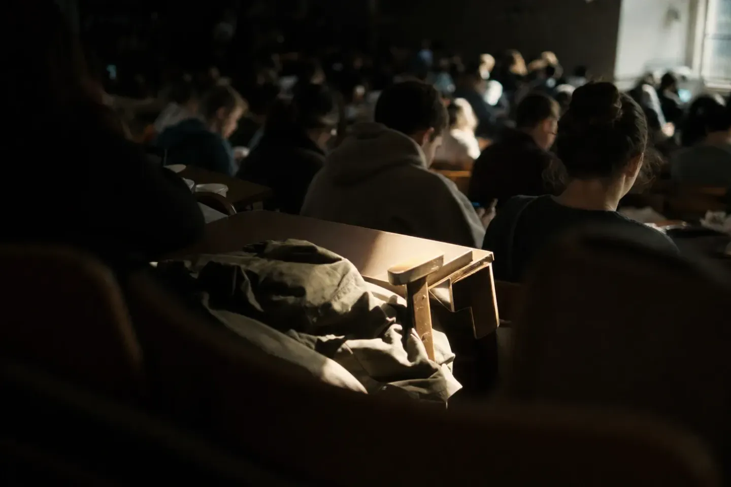 Un aula que se llena para una clase de las 8, vista desde las últimas filas a la altura de los respaldos: filas de estudiantes desenfocados acomodándose y, en primer plano medio, un asiento vacío con la tablita rebatible baja, alcanzado por una franja de luz cálida y dorada que entra por una ventana lateral; encima de la tablita, una campera arrugada, dejada de cualquier manera