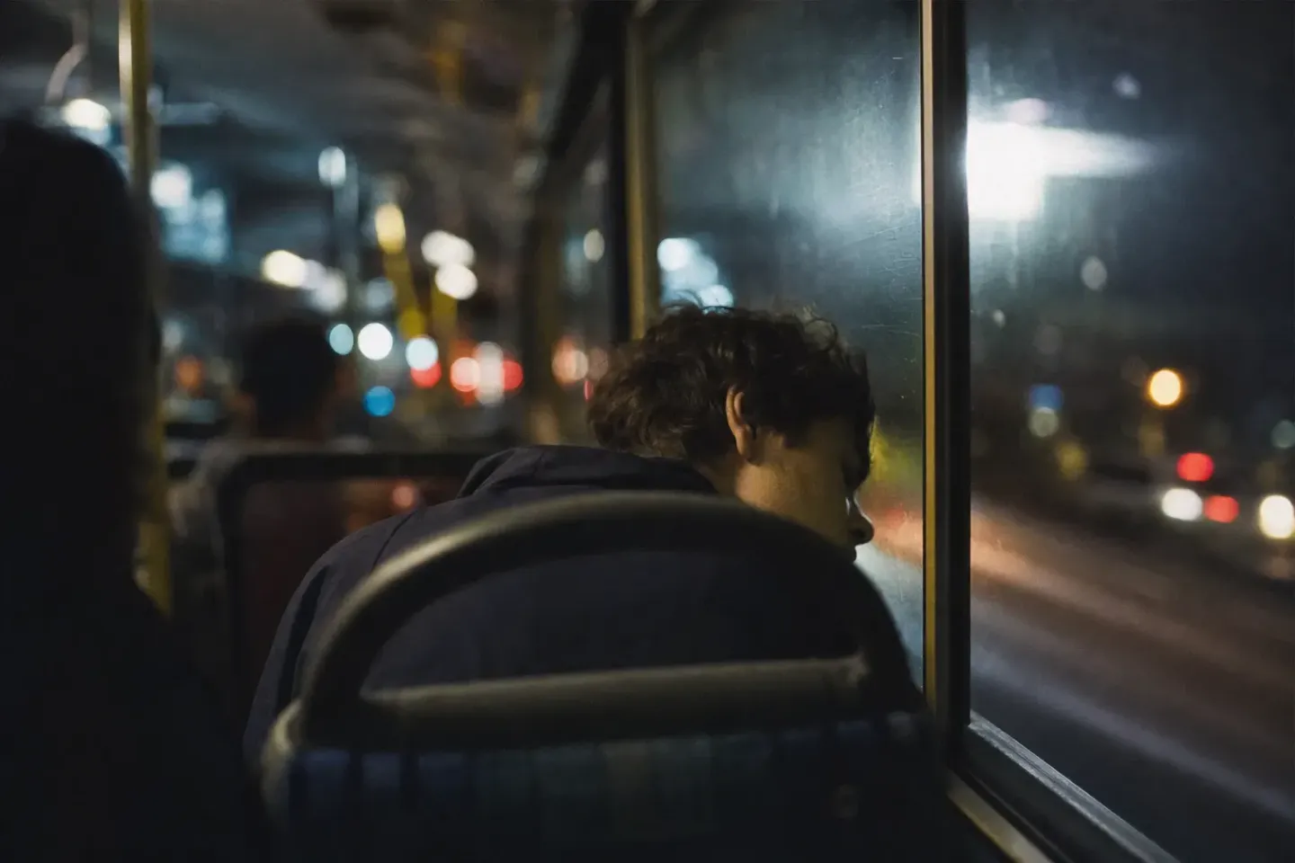 Passageiro dormindo sentado num ônibus à noite, a testa encostada na janela, com as luzes da cidade passando riscadas do lado de fora
