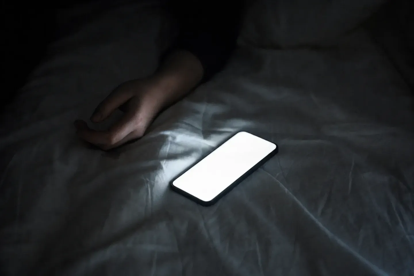 불 꺼진 방, 구겨진 침구 위에 화면을 위로 하고 놓인 휴대폰을 비스듬히 내려다본 사진. 화면은 하얗게 번져 아무것도 읽히지 않고, 그 옆으로 이불 밖에 나온 손이 힘없이 늘어져 있다
