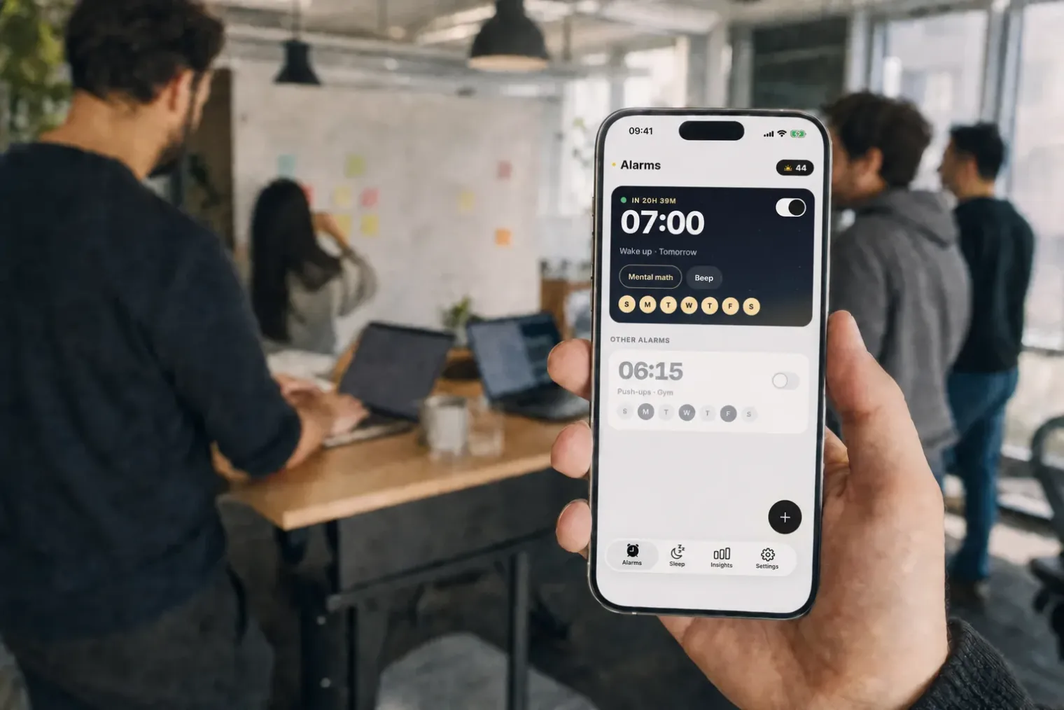 The Risly team at work in an open-plan office, one hand holding an iPhone with the Risly home screen.