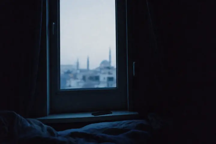Vue depuis un lit dans une chambre encore sombre : la fenêtre forme un rectangle pâle où l’on devine à peine des toits, un téléphone posé face contre l’appui