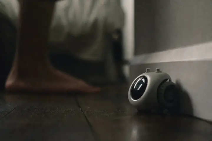 A small wheeled alarm clock jammed against a skirting board in a dark bedroom, seen from floor level, with a blurred bare foot behind it