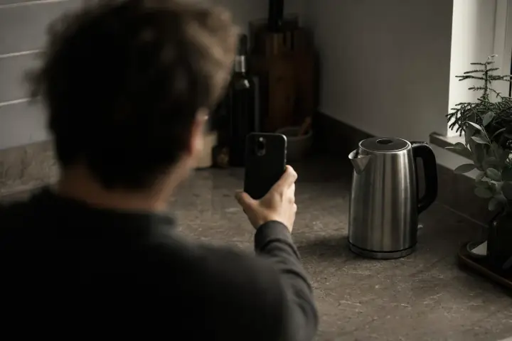 Seen over a blurred shoulder, someone points the back camera of a phone at a stainless-steel kettle standing in focus on a dim kitchen counter