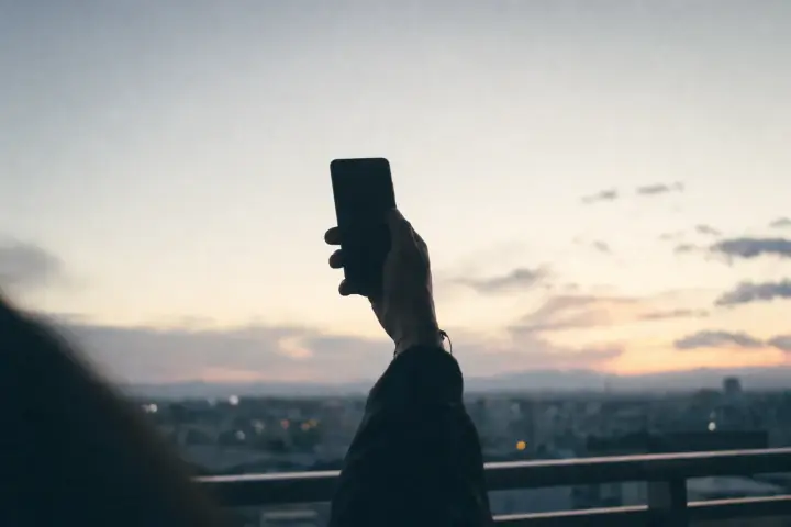 A silhouetted arm holding a phone up against a pale dawn sky, with a balcony rail and the lights of a waking city below, the screen turned away and unreadable