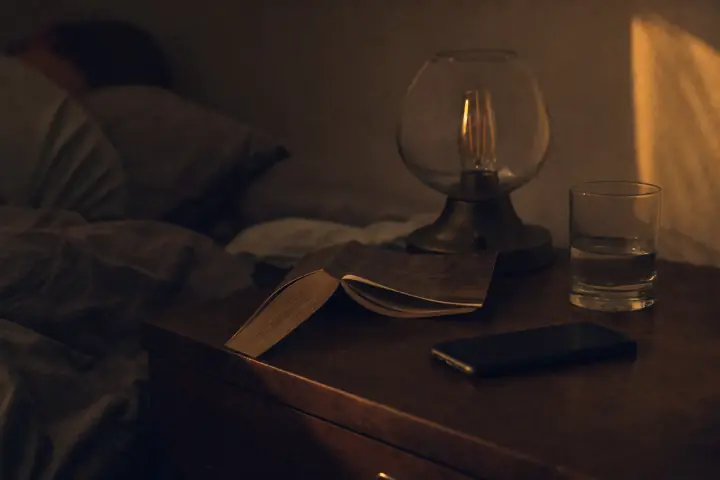 Une table de chevet la nuit : la lampe encore allumée, un livre de poche posé à plat ouvert, un verre d’eau, le téléphone repoussé, quelqu’un déjà endormi