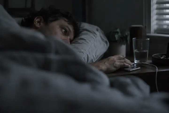 A man lying awake on his side before sunrise, seen at mattress height past the blurred hill of a duvet, one hand resting on a face-down iPhone on the nightstand next to a glass of water, gray dawn showing through the blinds